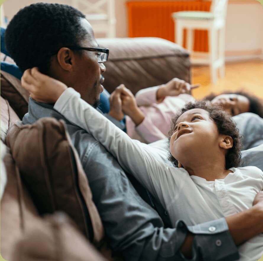 A father wearing glasses smiles down at his young child lying across his lap on a sofa, while a second child relaxes beside them