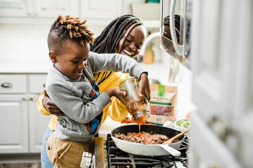 A happy mother and child cooking in the kitchen with the child being watched pouring sauce into a frying pan