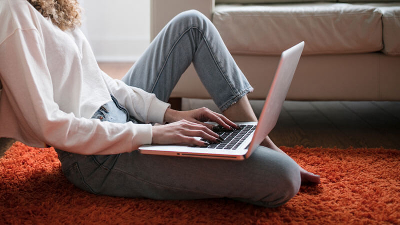 A lady sat on the floor whist working from her laptop which is balanced on her right leg.