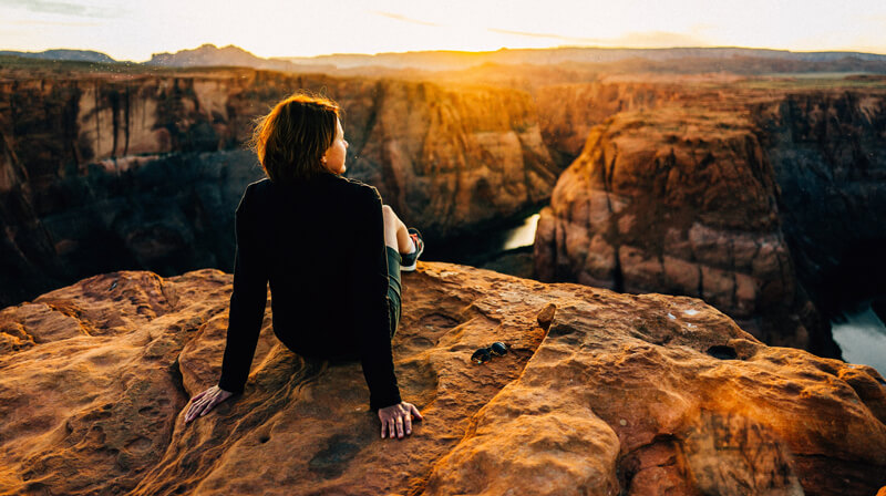 A woman sat on a rocky cliff overlooking the beautiful cavernous terrain with the golden sun setting in the background.