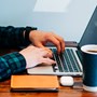 A person sat at a desk typing on their laptop with a coffee a notebook.