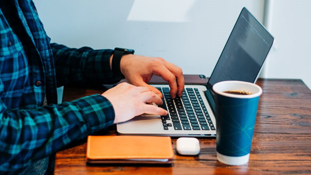 A person sat at a desk typing on their laptop with a coffee a notebook.