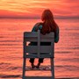 A woman sitting on a white chair looking out to sea during sundown. The sky is a picturesque deep orange and yellow. 