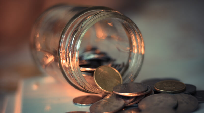 A glass money jar tipped over with a handful of copper coins spilling onto a countertop.
