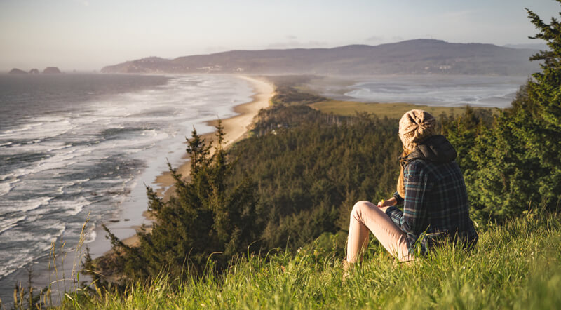 A lady sitting on a grassy hill overlooking the picturesque view of the blue sea meeting with the sandy beach.