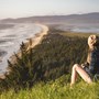 A lady sitting on a grassy hill overlooking the picturesque view of the blue sea meeting with the sandy beach.