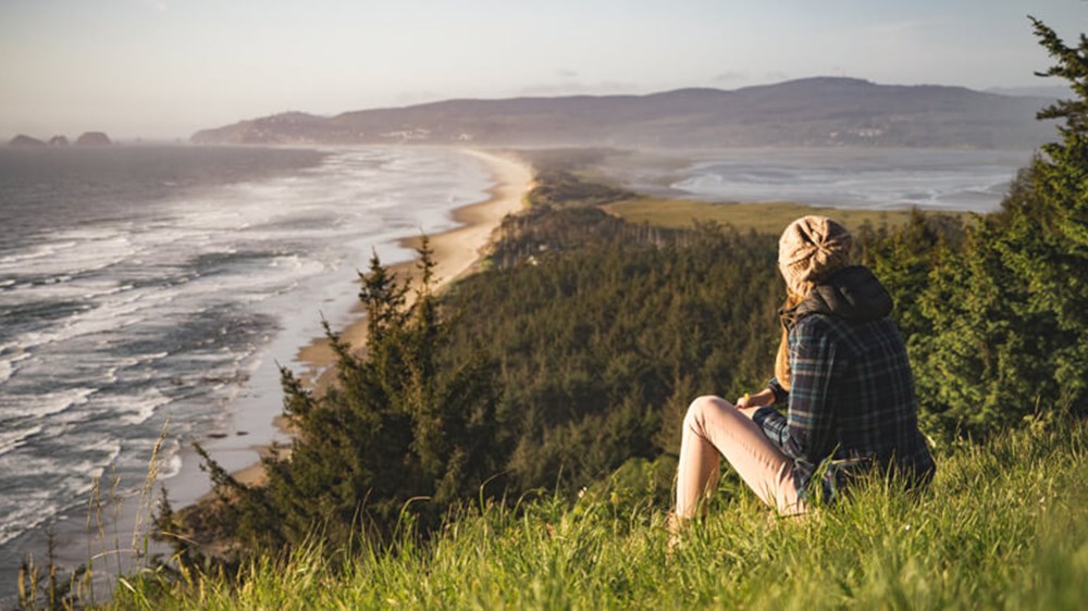 A lady sitting on a grassy hill overlooking the picturesque view of the blue sea meeting with the sandy beach.