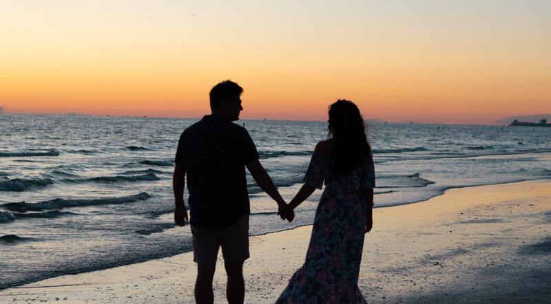 A couple walking along a beach at sunset holding hands.