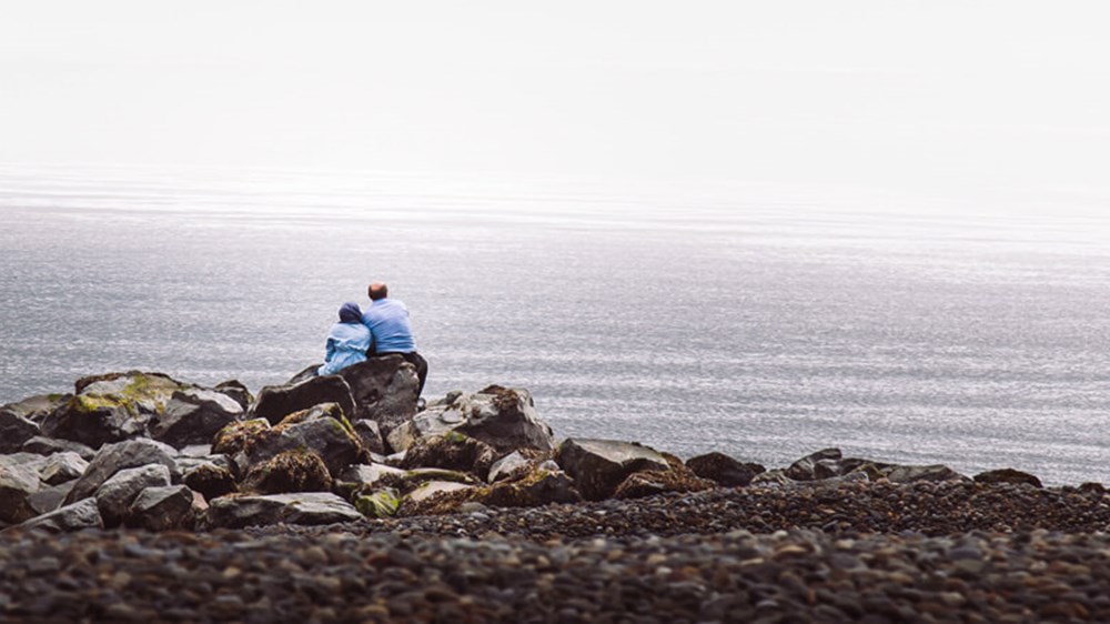 A couple sitting on a pile of big rocks overlooking a scenic view of the sea.
