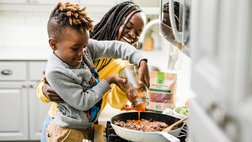 A mother and child cooking spaghetti and smiling. The child is pouring sauce into the frying pan while the parent supervises.