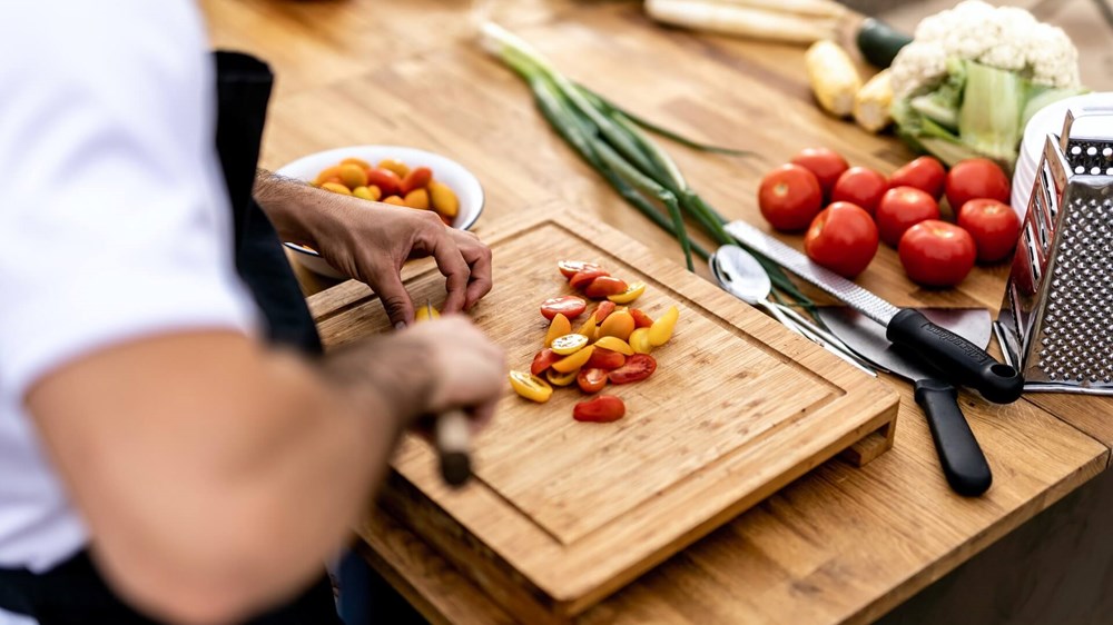 A chef in a white shirt and black apron cutting baby plum tomatoes on a wooden chopping board with an assortment of vegetables next to it | wealthify.com