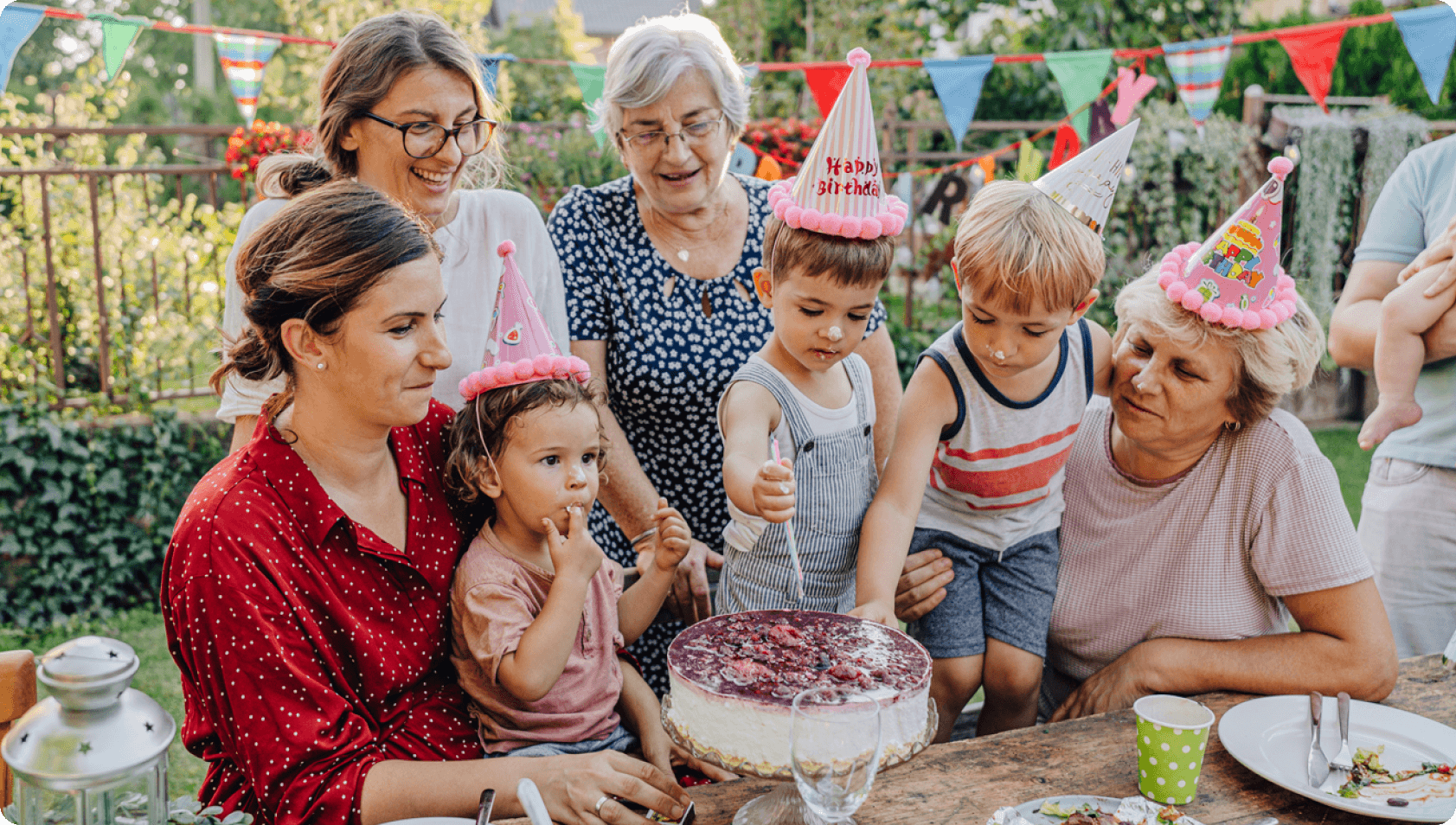 A family of 7 gathered around a child cutting on a cake on their birthday. 4 of them are wearing party hats.