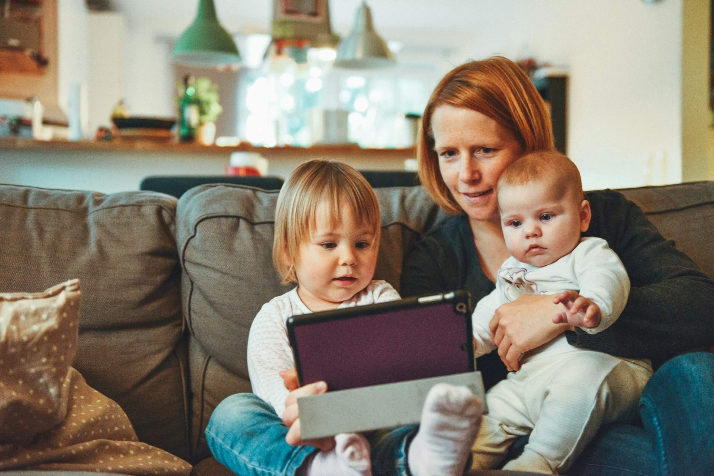 a mother sat on the sofa with her two children in her lap. She is showing the older child how to use a computer tablet.