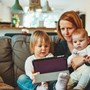 a mother sat on the sofa with her two children in her lap. She is showing the older child how to use a computer tablet.