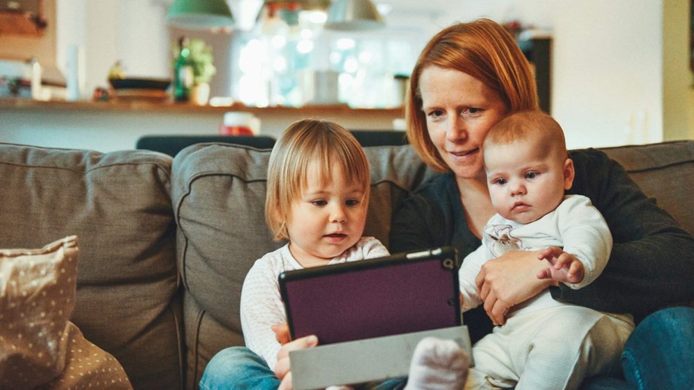 a mother sat on the sofa with her two children in her lap. She is showing the older child how to use a computer tablet.