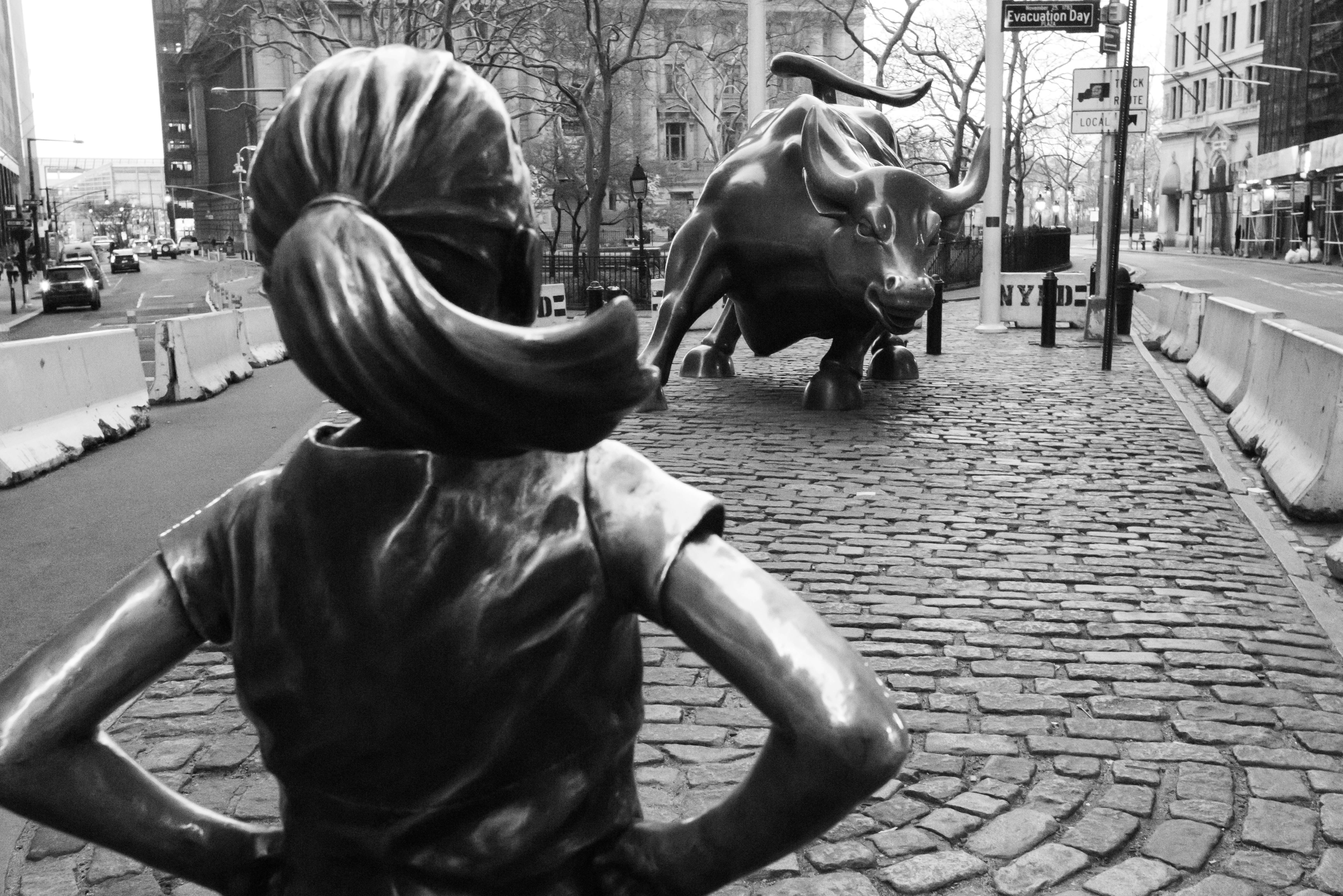 'Grayscale photo of fearless girl statue on Wall Street staring down a bull statue'