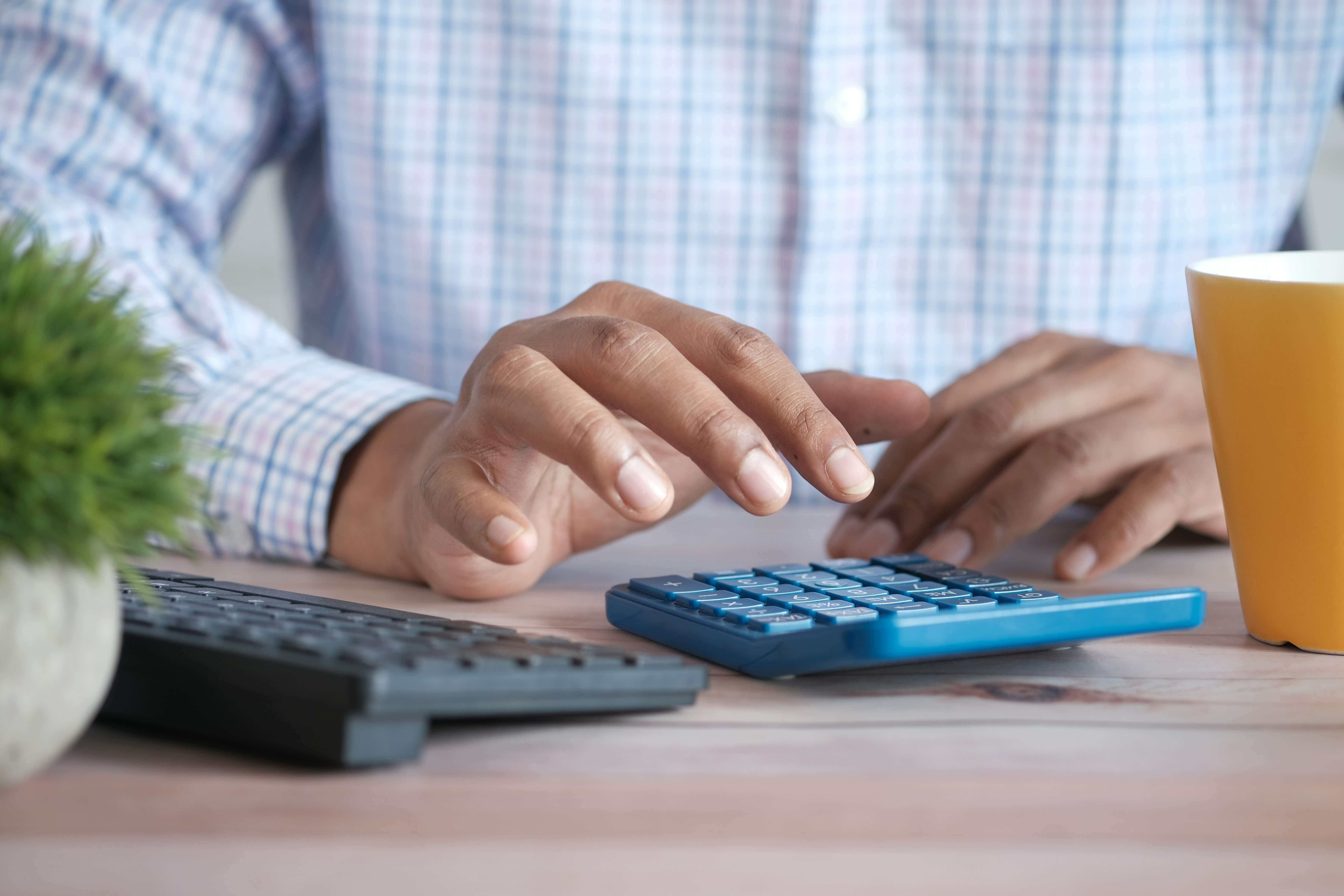 Close up photo of a man's hands using a calculator. There's a yellow coffee cup on the table as well as a small plant and computer keyboard.