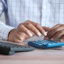 Close up photo of a man's hands using a calculator. There's a yellow coffee cup on the table as well as a small plant and computer keyboard.