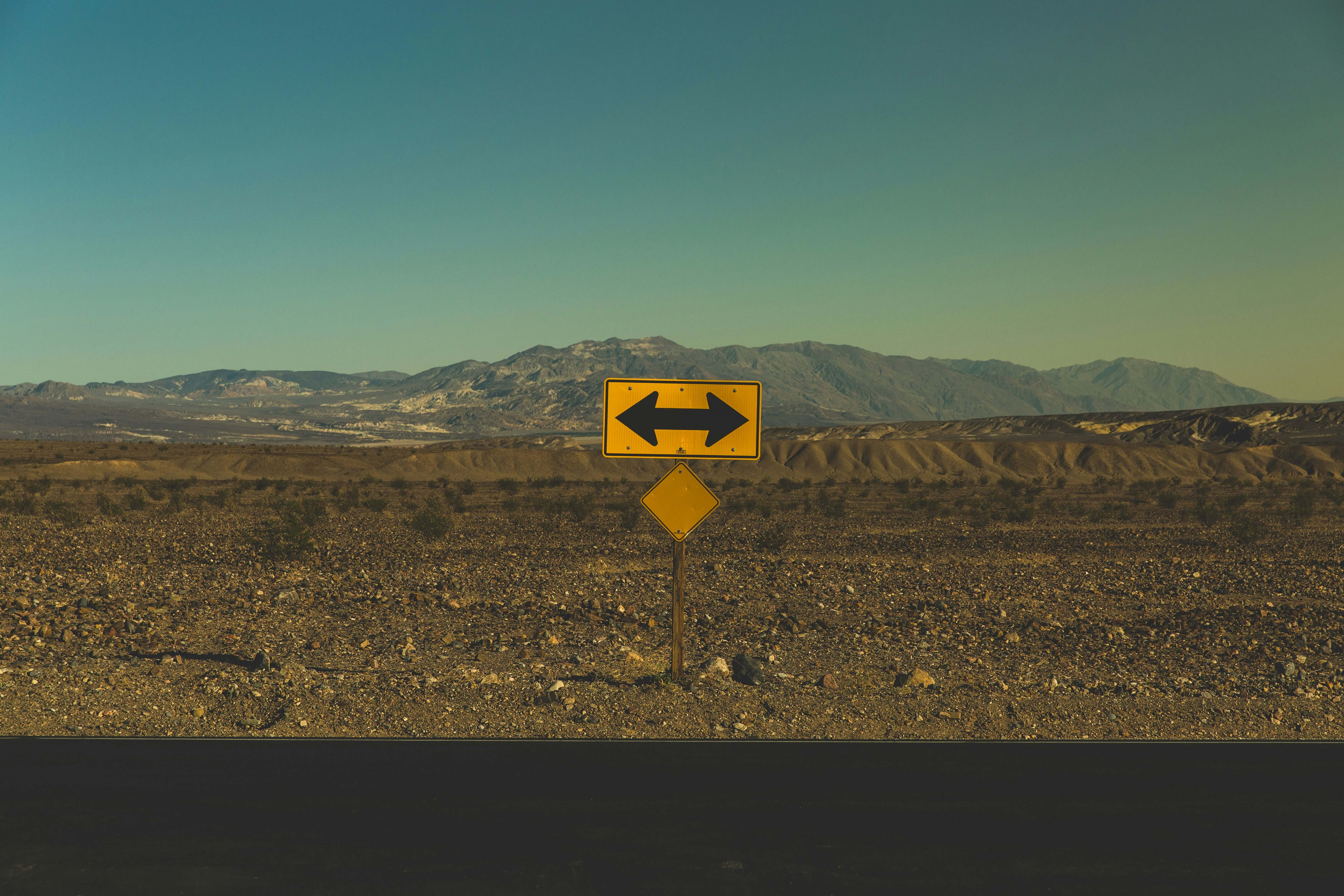 A landscape of a desert with a yellow road sign with arrows pointing both left and right.