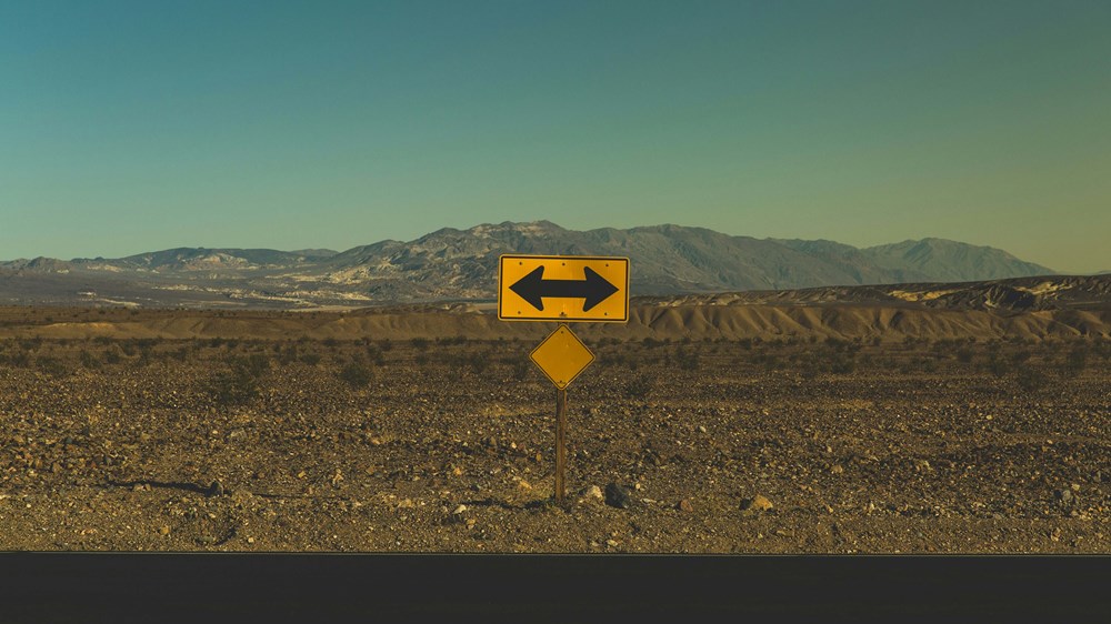 A landscape of a desert with a yellow road sign with arrows pointing both left and right.