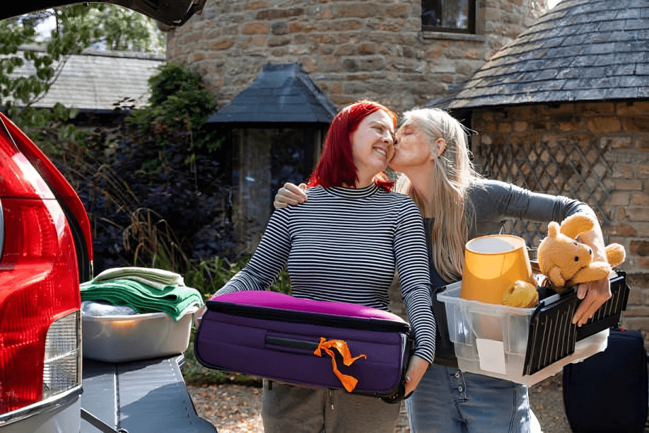 A mother kissing her teenage child goodbye as she's moving out. The teenager is carrying a suitcase and the mother is carrying a plastic box with a teddy bear and a lamp inside.