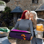 A mother kissing her teenage child goodbye as she's moving out. The teenager is carrying a suitcase and the mother is carrying a plastic box with a teddy bear and a lamp inside.