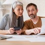 A middle aged couple sat at a desk looking through letters while drinking a cup of tea