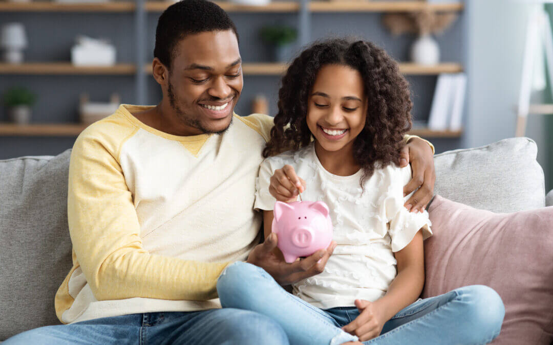 A dad sitting on the sofa with his child. The dad is holding a piggy bank for the child to put money in.