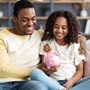 A dad sitting on the sofa with his child. The dad is holding a piggy bank for the child to put money in.