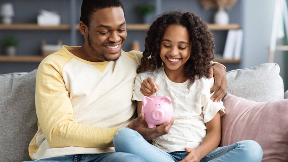 A dad sitting on the sofa with his child. The dad is holding a piggy bank for the child to put money in.