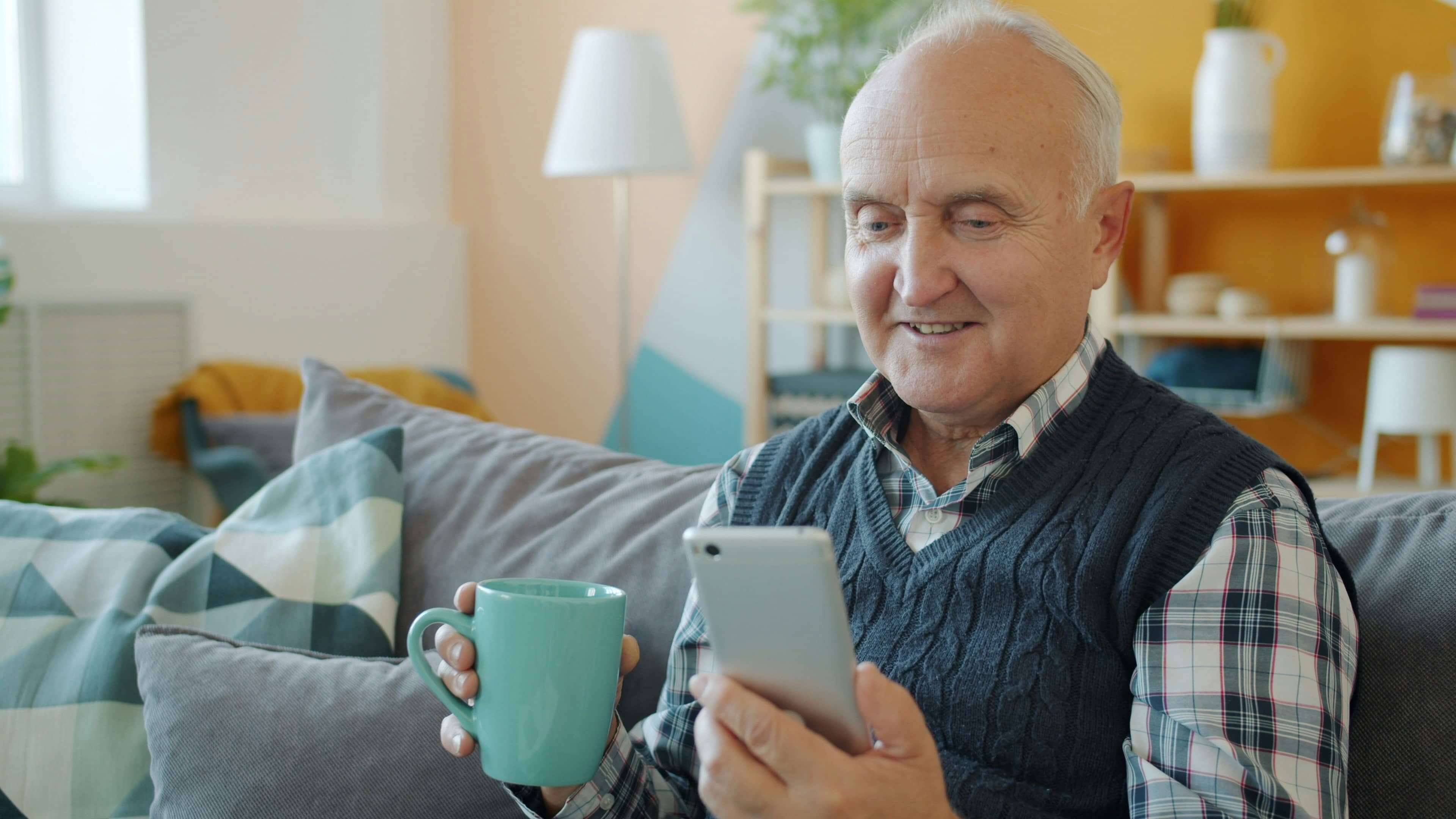 An older gentleman sat on a sofa with a mug in one hand and a smart phone in the other.