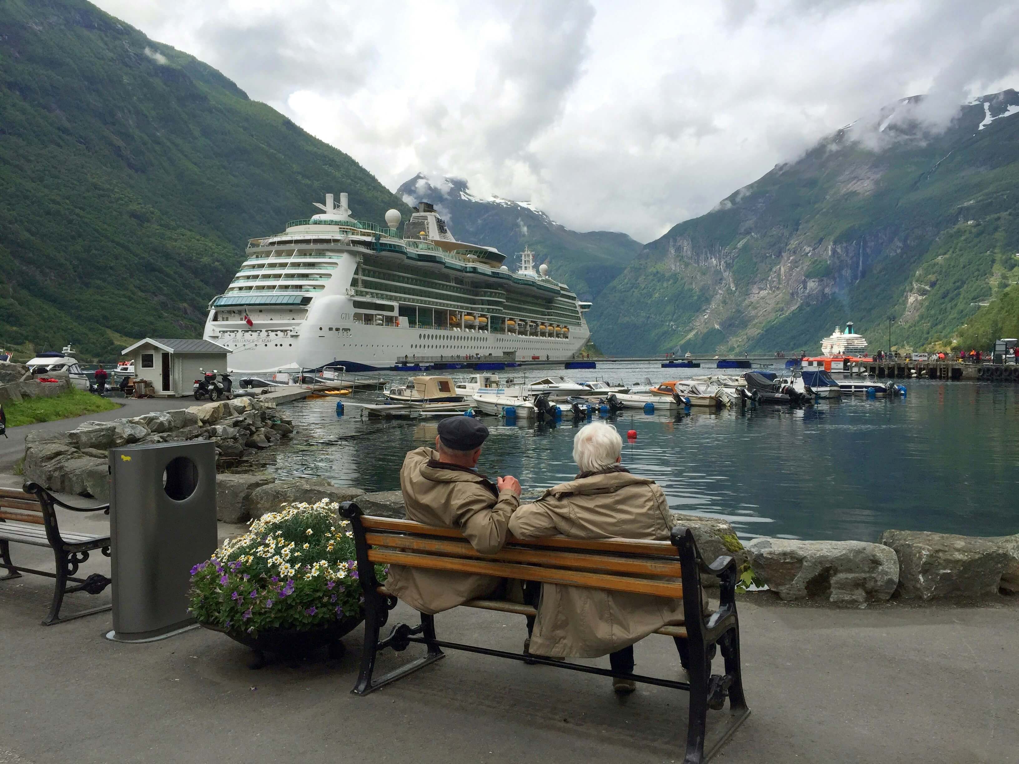 Two pensioners sat on a bench watching a cruise ship that's docked on a lake. The lake resides between two grassy hills.