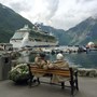 Two pensioners sat on a bench watching a cruise ship that's docked on a lake. The lake resides between two grassy hills. 