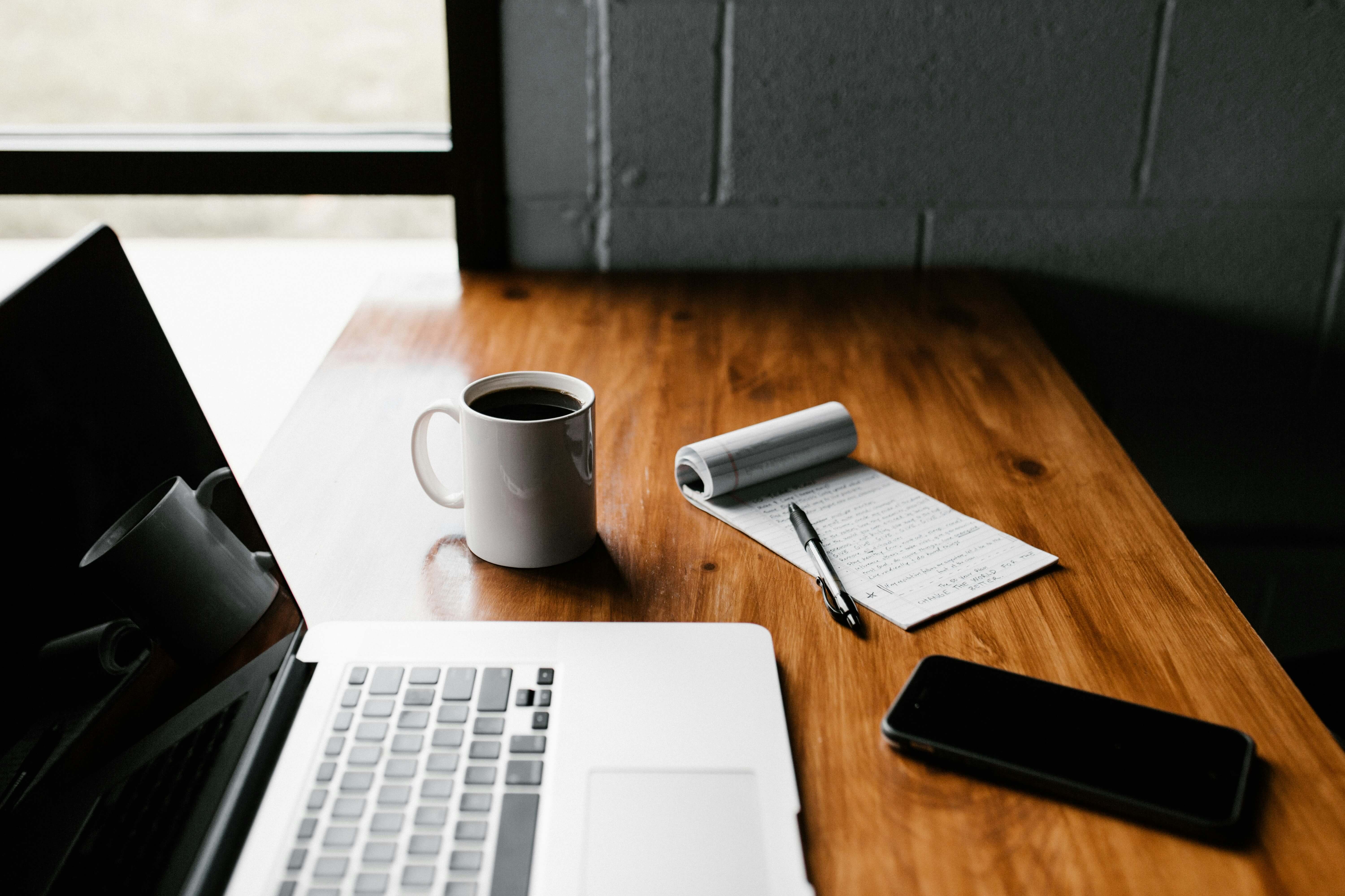 A wooden desk in front of a window with a laptop, cup of coffee, and note pad on it